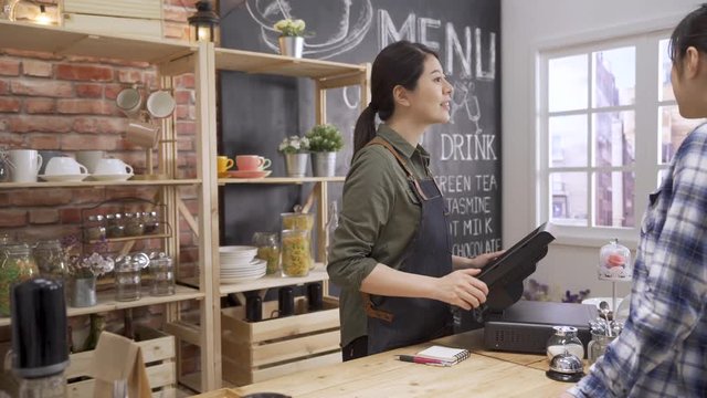 asian female customer ordering at counter in coffee shop. friendly woman waitress finger touching 3d rendering point of sale system for store management. two ladies looking at menu in cafe service