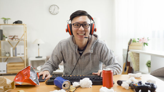 Front View Of Playful Young College Boy Playing Video Games In Messy Apartment On Summer Break At Home. Homebody Guy Smiling Looking At Camera As Computer Screen Wearing Headphones Excited Emotion.