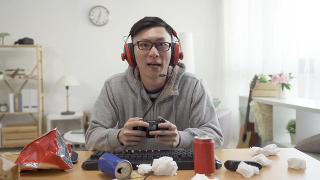 asian weird otaku playing video online anime game emotionally. young college boy student on summer break at home wear headphones holding controller staring at computer screen face camera intense.