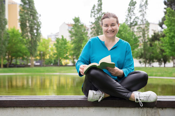 Obraz premium Happy middle aged student enjoying studying outdoors. Cheerful Caucasian lady sitting with legs crossed on park bench, holding book and smiling at camera. Studying in any age concept