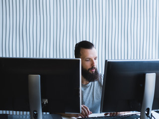 Computer programming. Bearded software developer using two monitors, working on task at office....