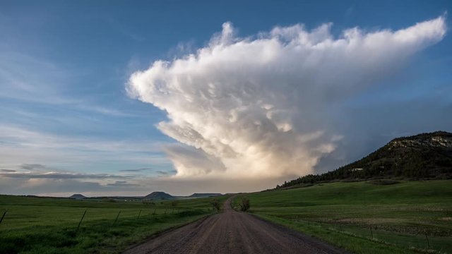 Time lapse of tall storm cell moving through the sky past Bald Mountain as dirt road head toward it disappearing in the distance.