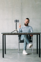 Social vlog. Bearded man using smartphone on tripod, greeting subscribers with peace gesture. Copy space.