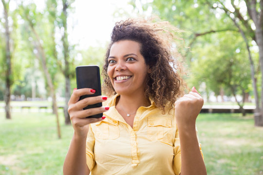 Cheerful Woman Reading News On Smartphone In City Park. Young Lady Pumping Fist And Standing With Blurred Green Trees And Lawn In Background. Success Concept. Front View.