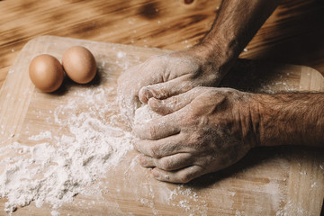 Beautiful and strong men's hands knead the dough from which they will then make bread, pasta or pizza. Next to the chicken egg. The background is dark.