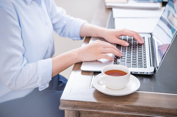Business concept. Woman in blue shirt typing on computer with coffee on office table, backlighting, sun glare effect, close up, side view, copy space