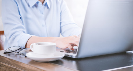 Business concept. Woman in blue shirt typing on computer with coffee on office table, backlighting, sun glare effect, close up, side view, copy space