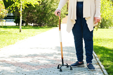 Senior disabled caucasian woman hands on cane outside nursing home park. Close up of elderly lady holding a walking stick outdoors of healthcare facility on the sunny day.