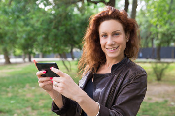 Smiling pretty lady browsing on smartphone in city park. Young woman looking at camera, holding gadget and standing with blurred green trees in background. Communication concept. Front view.