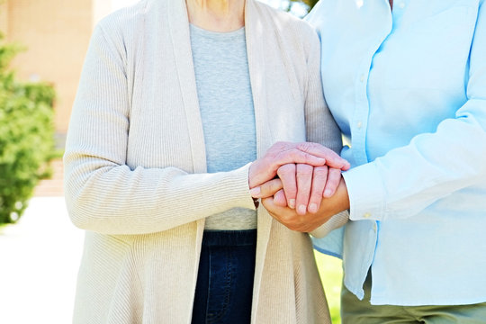 Mature Female In Elderly Care Facility Yard Gets Help From Hospital Personnel Nurse. Senior Woman, Wrinkled Skin & Hands Of Her Care Giver. Grandmother On Park Walk. Background, Copy Space, Close Up.