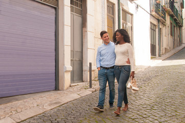 Obraz na płótnie Canvas Happy interracial couple enjoying long walk through old city. Carefree Afro American barefooted girl carrying her sandals and embracing her Caucasian boyfriend. Uncomfortable shoes concept