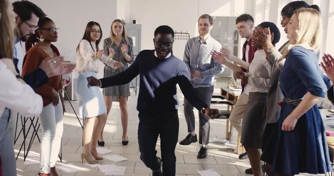 Fun happy young African businessman doing silly winner dance celebrating success with colleagues at workplace party.
