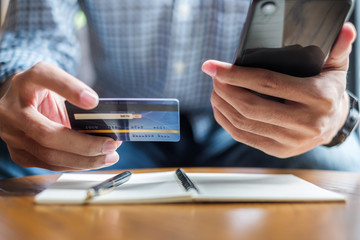 Young casual Business man writing something on notebook while holding credit card and using touchscreen smartphone in the cafe. business, lifestyle, technology, ecommerce and online payment concept