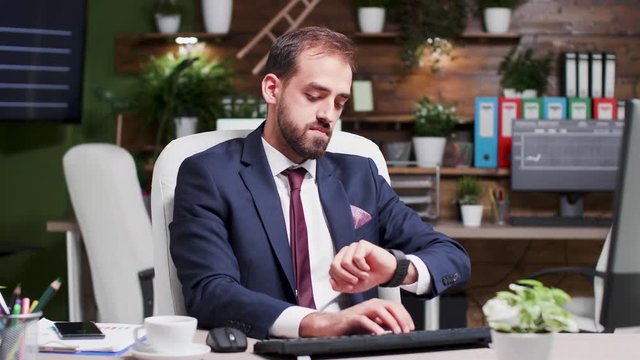 Businessman working extra hours in the week end looks at the time on his watch. Cozy and modern looking office in the background