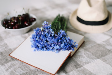 Flat lay of bouquet of cornflowers, straw hat and a book. Summer background