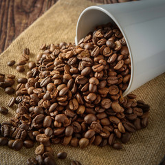 Coffee beans overflowing from a white coffee cup placed on a sackcloth and a black background.