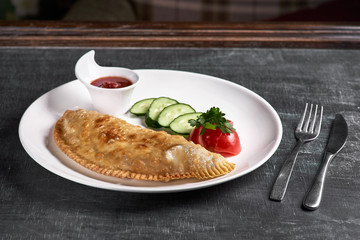 Uzbek eastern Tatar cuisine, cheburek with meat and suluguni cheese in a white plate with vegetables and greens on a dark wooden background.