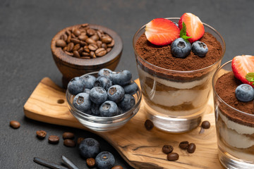 Classic tiramisu dessert with blueberries and strawberries in a glass on dark concrete background