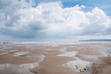 minimalist image of a flat sandy beach at Filey, North Yorkshire, with numerous puddles left behind by a low tide, with blue sky and white fluffy clouds reflecting, Bempton Cliffs in the background