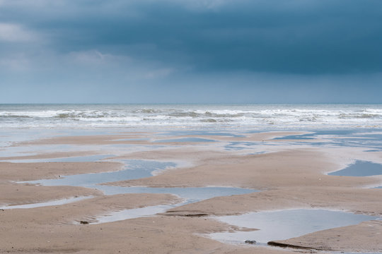 minimalist image of a flat sandy beach at Filey, North Yorkshire, with numerous puddles left behind by a low tide, with low, heavy dark grey clouds reflecting in the puddles