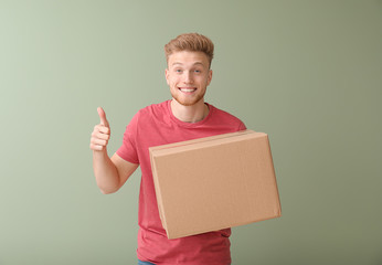 Young man with cardboard box showing thumb-up on color background