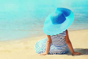 Fototapeta na wymiar Summer holidays and vacation concept - little girl in striped dress, straw hat enjoying sitting on sand beach near sea