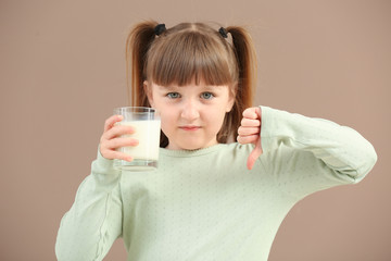 Cute little girl with glass of milk showing thumb-down on color background