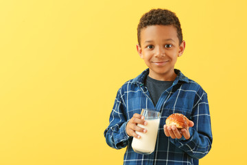 Cute African-American boy with glass of milk and bun on color background