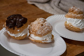 powdered sugar and shiny chocolate glased cream puffs with a swirl of white whipped cream on a paper plate