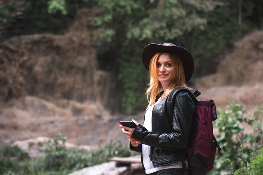 Female tourist use smartphone, with backpack and cowboy hat looking at the river washed away the bridge, the crossing was impossible. Deep waterfall. Copy space. adventure concept. network enternet