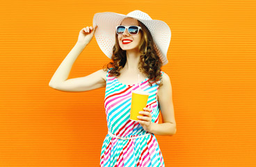 Portrait happy smiling young woman with cup of fruit juice in summer straw hat, colorful striped dress on orange wall background