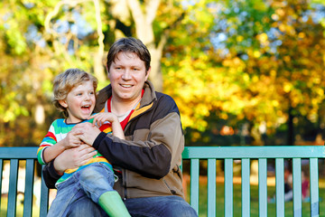 Little kid boy and young father sitting together in colorful clothing on bench in park. Cute healthy child and dad, middle-aged man having fun in autumn park on warm sunny day. Happy family of two.