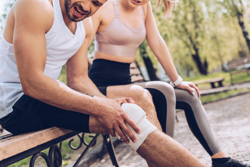 Obraz premium cropped shot of sportsman suffering from pain in injured knee while sitting on bench near young sportswoman