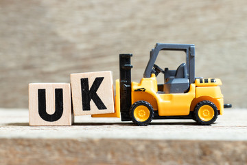 Toy forklift hold letter block k to complete word UK (abbreviation of united kingdom) on wood background