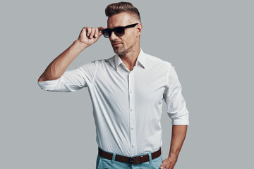 Modern macho. Handsome young man smiling and adjusting sunglasses while standing against grey background