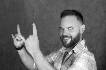 Black and white portrait of smiling bearded man showing rock gesture on gray background.