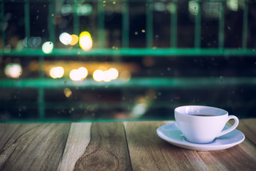 White cup of coffee on wooden table near windows in night bokeh, White cup of caffe americano on wooden table near windows in night bokeh.