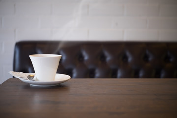 White cup of coffee on wooden table in the cafe, free space with old brown vintage sofa background.