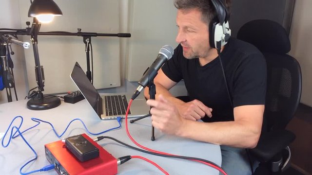 A podcaster or radio presenter recording in a podcast studio. He records his voice on a dynamic microphone and reviews and edits on a laptop.