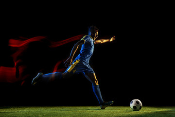 Young african-american male football or soccer player in sportwear and boots kicking ball for the goal in mixed light on dark background. Concept of healthy lifestyle, professional sport, hobby.