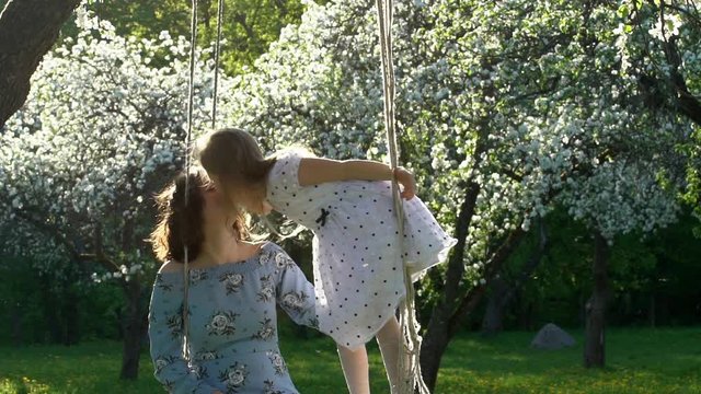 Lovely girl in dress kiss mother stand on swings in blossom garden. Slow motion
