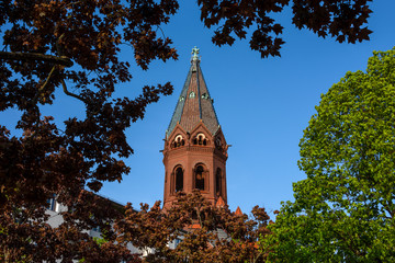 Fototapeta na wymiar Germany, Berlin, Kreuzberg, Bergmannstrasse: Steeple and part of the red brick facade of the church Passionskirche with green trees and blue sky in the background - concept religion architecture