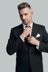 Getting ready. Handsome young man adjusting his necktie and smiling while standing against grey background