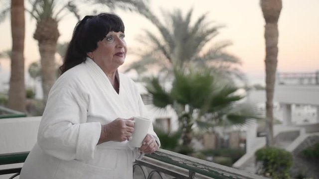 Caucasian senior woman in the morning drinking coffee on the hotel terrace in a white terry robe at a tropical resort. Against the backdrop of palm trees and the sea. Slow Motion, closeup.