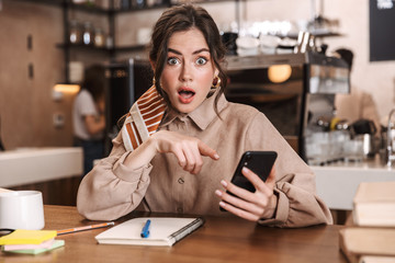 Shocked young beautiful woman using mobile phone indoors in cafe.