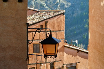 Elegant glass metal street lamp attached to a corner of a light brown house on a narrow rural street. Between the walls you can see the slope of the mountain. Albarracin village, Teruel, Aragon, Spain