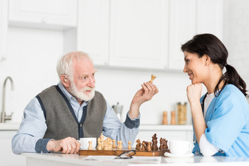 Nurse and grey haired man sitting on bright kitchen, and playing in chess