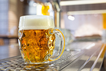 Freshly poured draft lager beer in a dimpled glass mug on stainless steel counter in a modern pub. Space for text. - obrazy, fototapety, plakaty