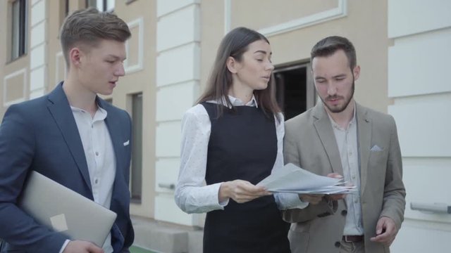 Young woman and two men in formal wear walking on terrace discussing project. The girl nervous, she throwing up papers and walking away. Concept of freelance, distant work. The guys working outdoors