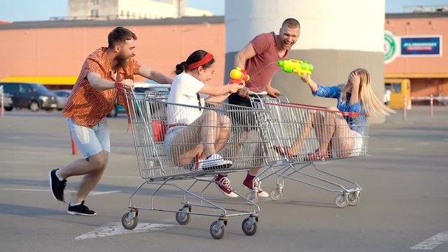 Young friends running with supermarket carts at the paking and playing water gun fight. Handheld shot.